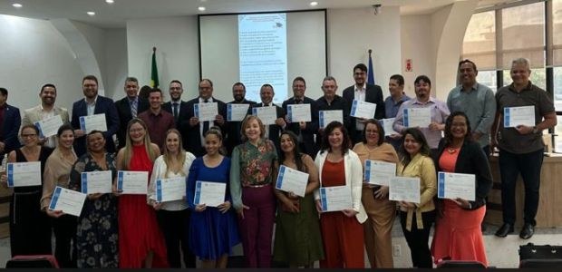 Na foto, várias pessoas, entre homens e mulheres estão em um auditório de paredes brancas. Elas ...