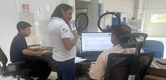 Na foto, 3 pessoas aparecem trabalhando. São 2 homens sentados em frente a tela de computadores ...