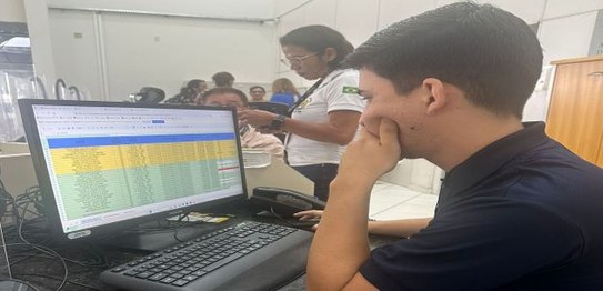 Na foto, 3 pessoas aparecem trabalhando. São 2 homens sentados em frente a tela de computadores ...