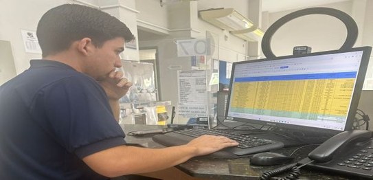 Na foto, 3 pessoas aparecem trabalhando. São 2 homens sentados em frente a tela de computadores ...