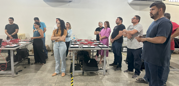 Na foto, várias pessoas adultas, entre homens e mulheres estão em uma grande sala de paredes bra...