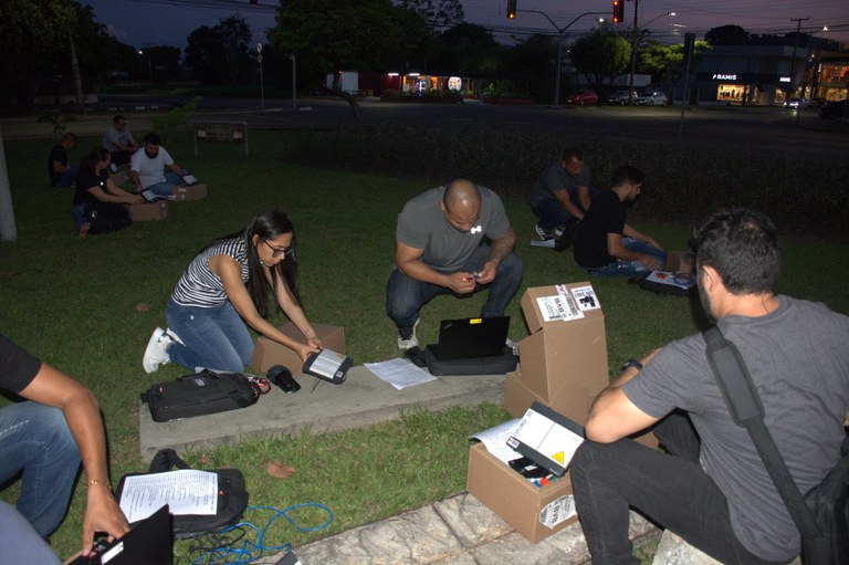 Várias pessoas adultas estão sentadas em um gramado. Eles utilizam vários notebooks e telefones ...