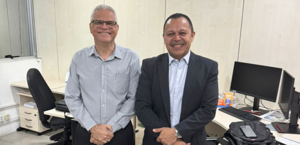 Na foto em uma sala de paredes brancas, 2 homens em pé. Por detrás deles, mesas, computadores e ...