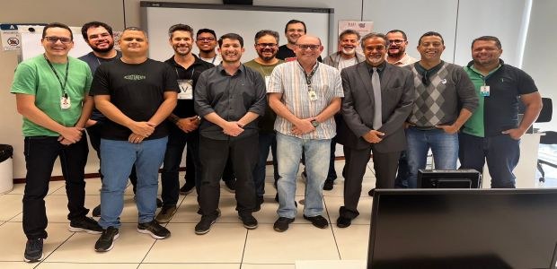 Na foto, em uma sala de videoconferência ampla e de paredes brancas, temos vários homens em pé, ...