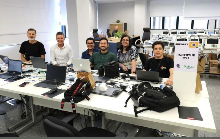 Na foto, 06 homens estão sentados na frente de uma bancada. Entre elas, 02 são mulheres e 04 são...